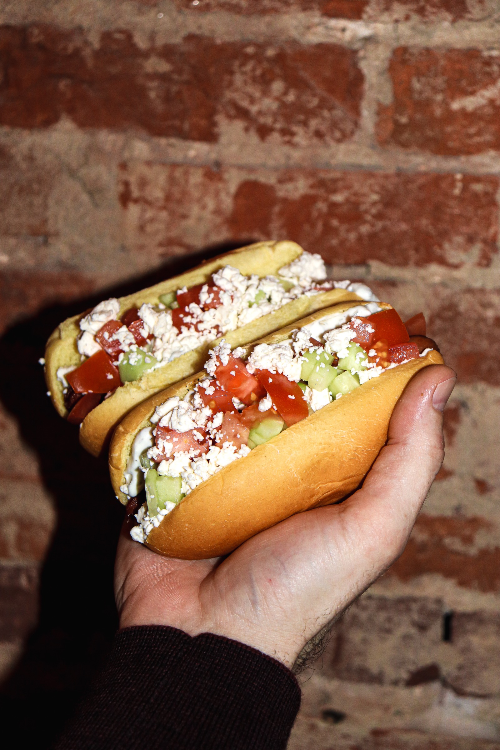 Two feta-and-tomato glizzies held against the brick wall at Jitters